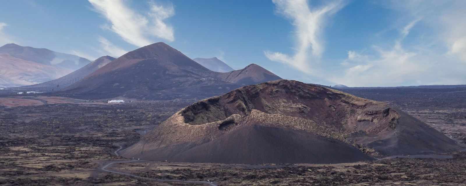 Volcanic landscape at Montaña Cuervo on Lanzarote, showing a wide trail curving toward a large crater surrounded by dark lava fields and distant volcanic hills under a bright blue sky.