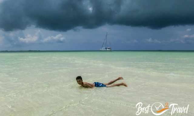 A boy laying in the turquois water of the lagoon and a catamaran in the back
