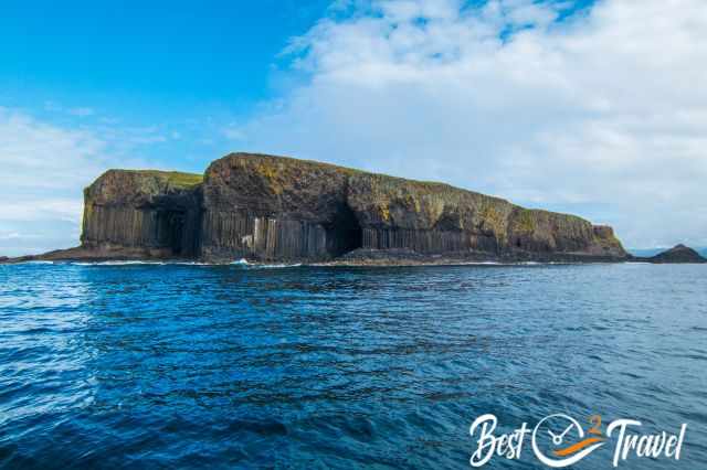 Basalt cliffs of the Isle of Staffa rising steeply from the deep blue sea, formed by tall hexagonal columns with sparse vegetation on top under a partly cloudy sky.