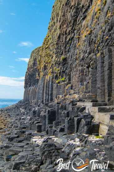 Basalt columns on the Isle of Staffa rising vertically from the rocky shoreline, covered with patches of green vegetation and lichen under a partly cloudy sky.