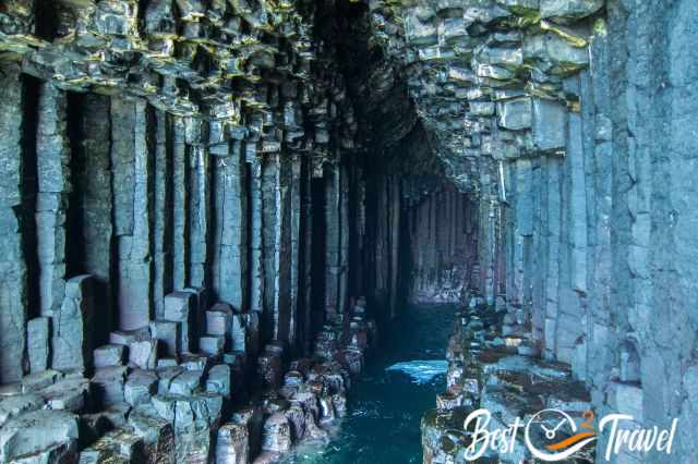 Interior of Fingal’s Cave with tall hexagonal basalt columns rising along the walls, opening into a narrow water-filled passage illuminated by natural light.