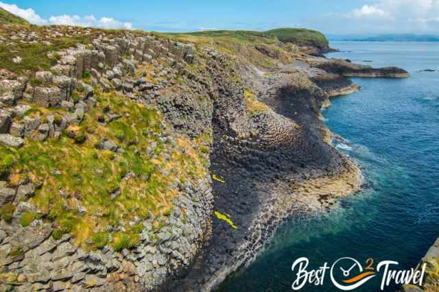 Terraced basalt columns on the Isle of Staffa descending toward clear blue water, with patches of grass and moss growing between the geometric rock formations.