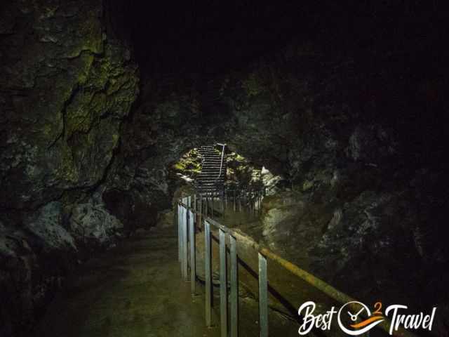 End section of Gruta das Torres lava tube guided tour with a metal walkway and staircase leading toward the illuminated exit, showing dark basalt walls and patches of moss.
