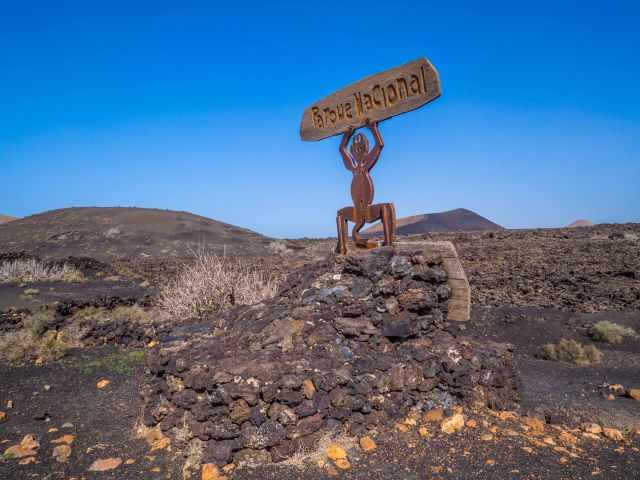 Metal Timanfaya entrance sculpture mounted on volcanic rocks, standing in a barren lava landscape with distant hills under a clear blue sky.
