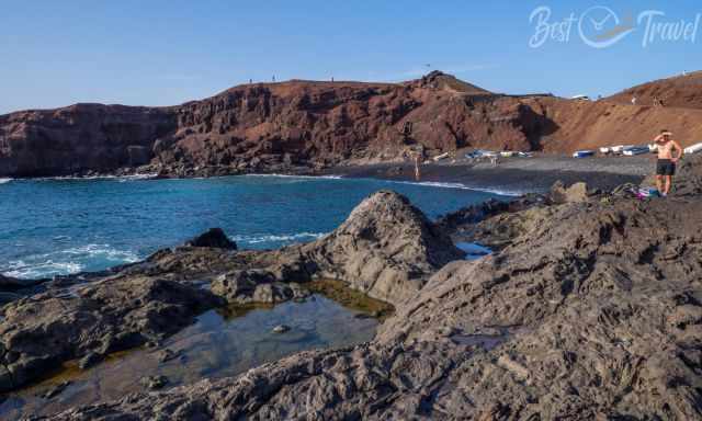 Volcanic bay at El Golfo on Lanzarote, with dark sand, small fishing boats, rugged lava rocks, and reddish crater cliffs.