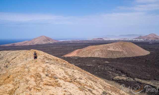 Hiker standing on the rocky summit ridge of Caldera Blanca, overlooking dark lava fields, volcanic cones, and the distant Atlantic coastline under a clear blue sky.