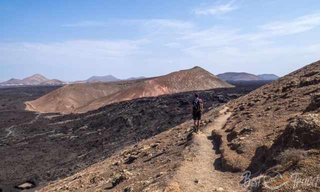 Hiker walking along the rocky descent trail from the summit of Caldera Blanca, surrounded by dark lava fields and reddish volcanic cones under a clear blue sky.