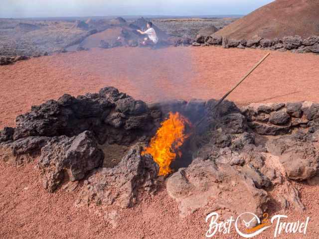 Flames rising from a geothermal vent in Timanfaya National Park, with a demonstration pole inserted into the pit and volcanic hills stretching across the barren landscape.