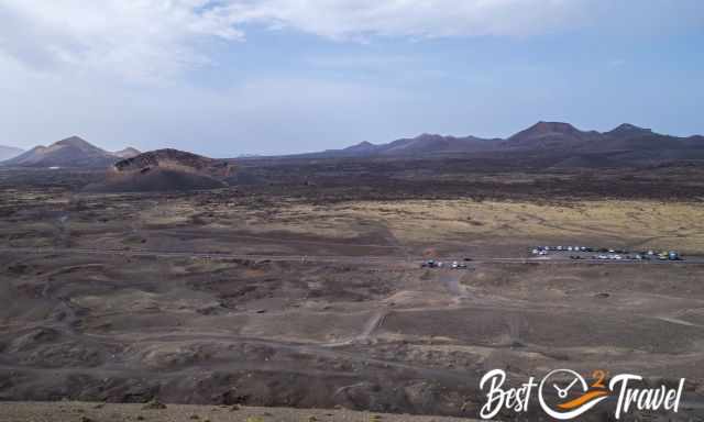 Wide volcanic landscape featuring Caldera de Los Cuervos and several surrounding volcanic cones in the natural park, with dark lava fields and winding paths under a partly cloudy sky.