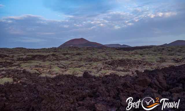 Volcanic landscape of Timanfaya seen from the El Golfo Trail, with dark lava fields, scattered vegetation, and distant volcanic cones under a partly cloudy sky.