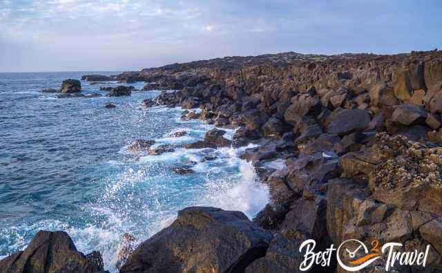 Waves crashing against sharp volcanic rocks along the El Golfo Trail, with dark lava cliffs and deep blue Atlantic water under a partly cloudy sky.