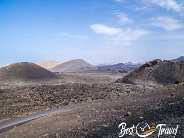 Wide volcanic landscape in Timanfaya National Park with rounded volcanic cones, dark lava fields, and a winding road leading through the barren terrain under a clear sky.