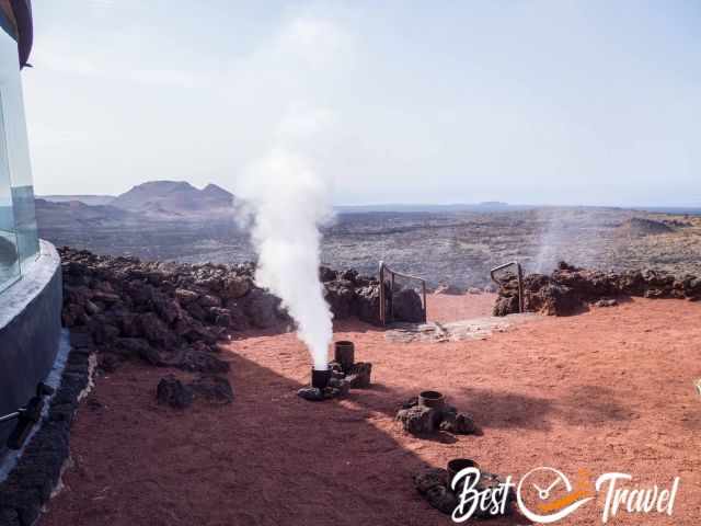 Geothermal vent releasing a plume of steam in Timanfaya National Park, surrounded by rugged volcanic rock, reddish soil, and distant lava hills under a clear sky.