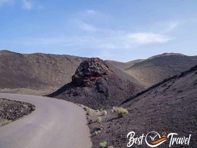 Winding road curving through Timanfaya National Park’s volcanic landscape, passing a mound of dark and reddish lava rock surrounded by barren hills