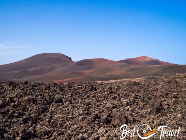 Rounded volcanic cones in Timanfaya National Park rising above dark lava fields, with rugged basalt rocks in the foreground and a clear blue sky overhead.