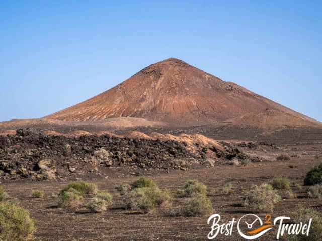 Reddish volcanic cone rising from dark lava fields in Timanfaya National Park, with scattered shrubs in the foreground.