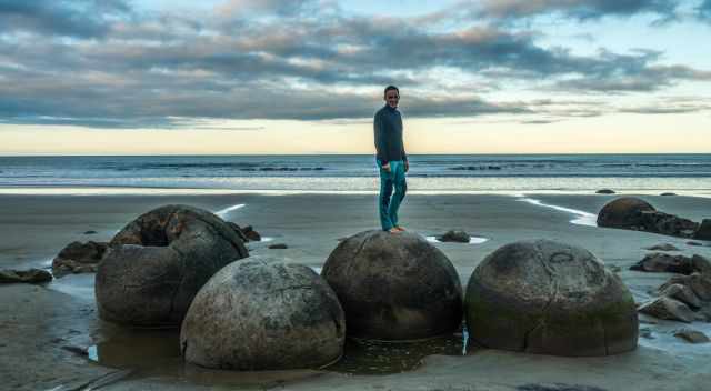 A man standing on a huge Moeraki Boulder with the sea in the back.