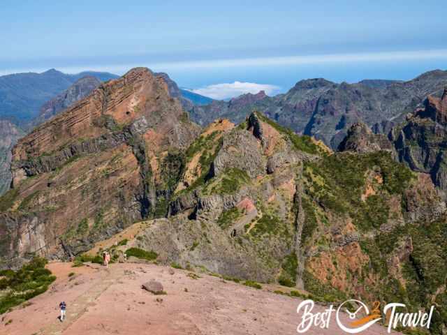 Hikers at the beginning of the PR1 trail from Pico do Arieiro, with the rugged peak of Pico das Torres rising above the dramatic mountain ridges.