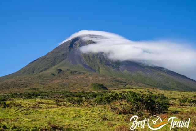 Pico Mountain rising above green volcanic terrain, partly wrapped in clouds near the summit on a clear day.