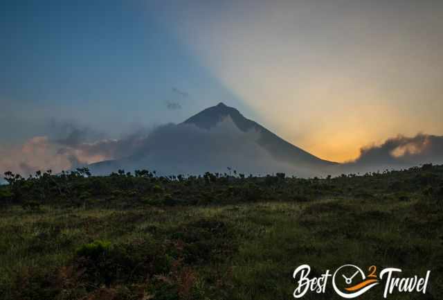 Pico Mountain at sunset, low clouds wrapping around the summit, and grassy volcanic terrain in the foreground.