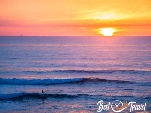 A surfer on a wave with an orange and pink sunset in the back