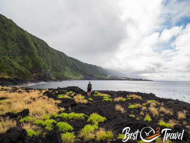 A man on a black lava field and sheer lush green cliffs