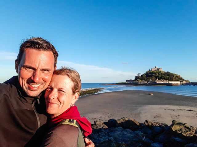Two people smiling with St Michael’s Mount in the background at sunset, standing on the beach with the island and castle softly lit by golden evening light.