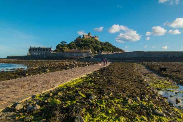 St Michael’s Mount in the evening at low tide with people walking the exposed stone causeway, seaweed-covered rocks in the foreground, and the castle rising under a bright sky.