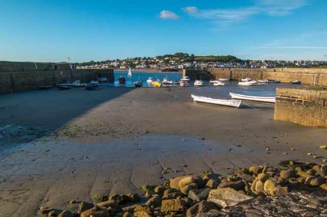 Small harbour at St Michael’s Mount at low tide with boats resting on the sand and a view across the water toward Marazion’s hillside houses.