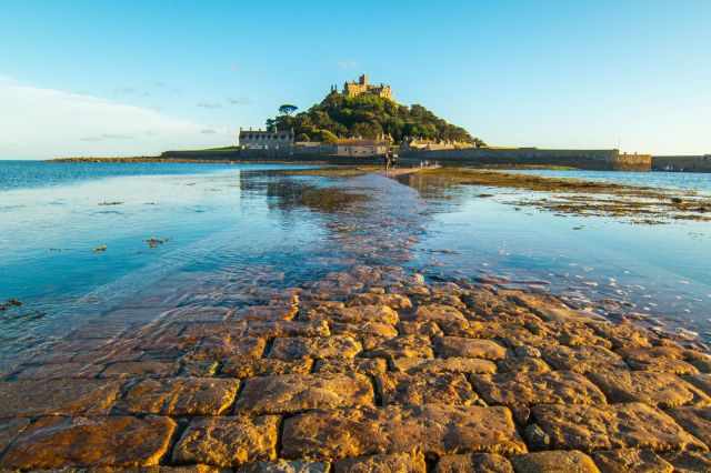 St Michael’s Mount at low tide in warm evening light, with the stone causeway exposed and shallow water reflecting the golden sky.