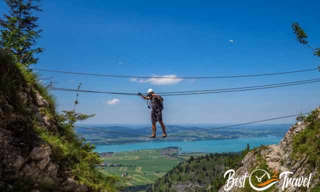 A climber on a rope bridge with two paragliders in the back